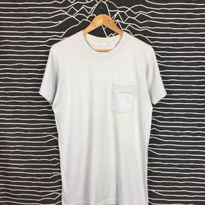 Könnte beinhalten: Ein schlichtes weißes T-Shirt mit einer einzigen Tasche auf der linken Seite. Das Shirt hängt an einem Holzbügel vor einem schwarz-weiß gestreiften Hintergrund.