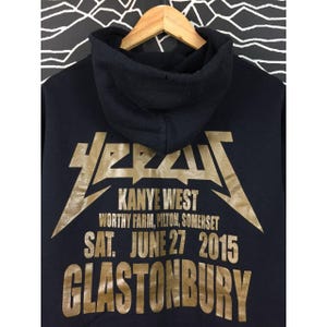 May include: Black hoodie with gold lettering. The text reads "Kanye West" and "Glastonbury" with the date "June 27 2015". The hoodie has a hood and is hanging on a wooden hanger.