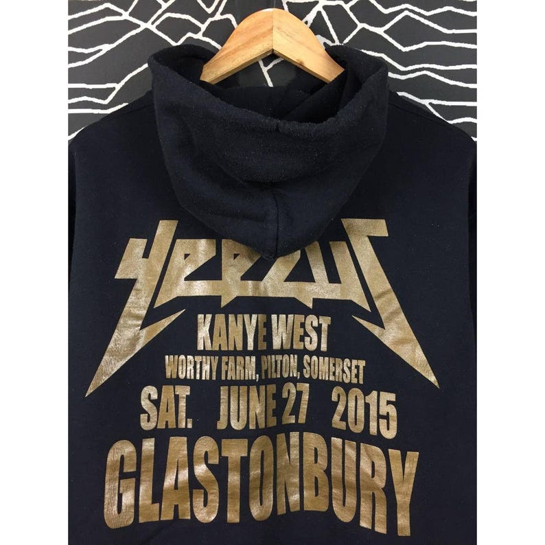 May include: Black hoodie with gold lettering. The text reads "Kanye West" and "Glastonbury" with the date "June 27 2015". The hoodie has a hood and is hanging on a wooden hanger.