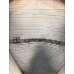 May include: A white button-down shirt featuring a subtle pattern of thin, horizontal light blue and yellow pinstripes. A rectangular fabric label is stitched at the collar, displaying the text "courr&egrave;ges homme PARIS" and a logo.
