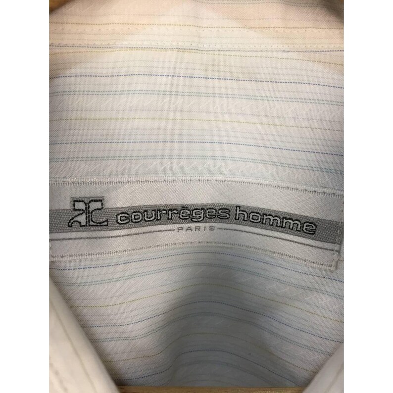 May include: A white button-down shirt featuring a subtle pattern of thin, horizontal light blue and yellow pinstripes. A rectangular fabric label is stitched at the collar, displaying the text "courr&egrave;ges homme PARIS" and a logo.