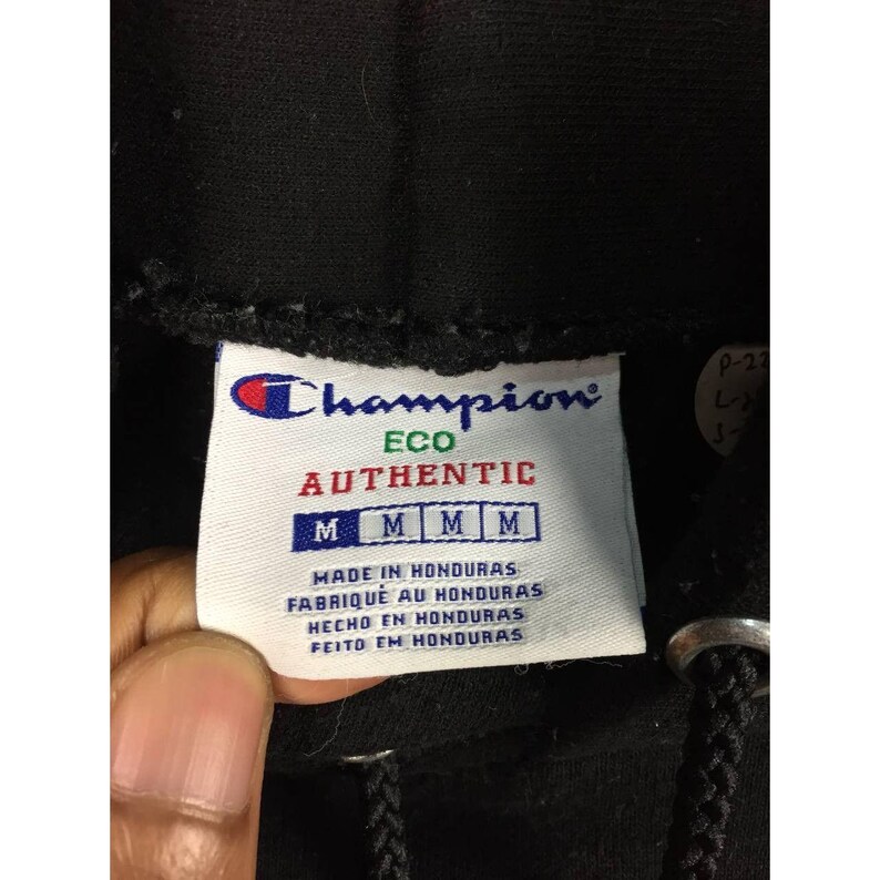 May include: Close-up of a Champion brand tag. The tag is white with the Champion logo in blue and red. The word "AUTHENTIC" is in red. The tag also has the text "MADE IN HONDURAS" and four "M" size indicators.