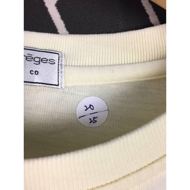 Op de afbeelding: Close-up van een lichtgele t-shirtkraag met een label met de tekst "ēges co". Een kleine, ronde sticker met de nummers "20" boven "25" is zichtbaar. Het shirt hangt aan een houten hanger.