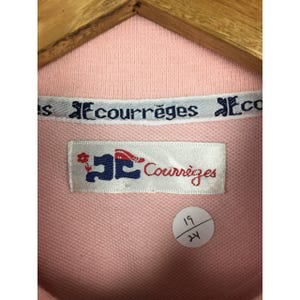 May include: Close-up of a light pink polo shirt with a white collar. The collar features the text "Courr&egrave;ges" in a repeating pattern. A label with the brand name and logo is visible. A small sticker with the numbers "19" and "24" is also present.