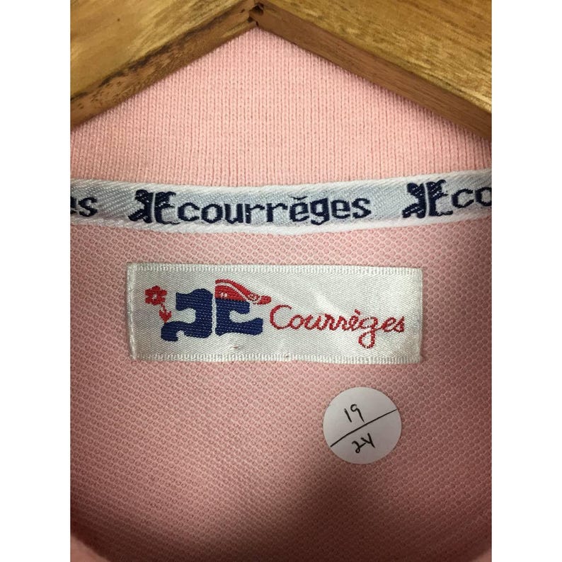 May include: Close-up of a light pink polo shirt with a white collar. The collar features the text "Courr&egrave;ges" in a repeating pattern. A label with the brand name and logo is visible. A small sticker with the numbers "19" and "24" is also present.