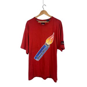 Puede incluir: Camiseta roja de manga corta con un gráfico de un encendedor azul con una llama en ángulo diagonal. El texto "SATURDAY NIGHT" y "HAPPY DAZE" está impreso en el encendedor.