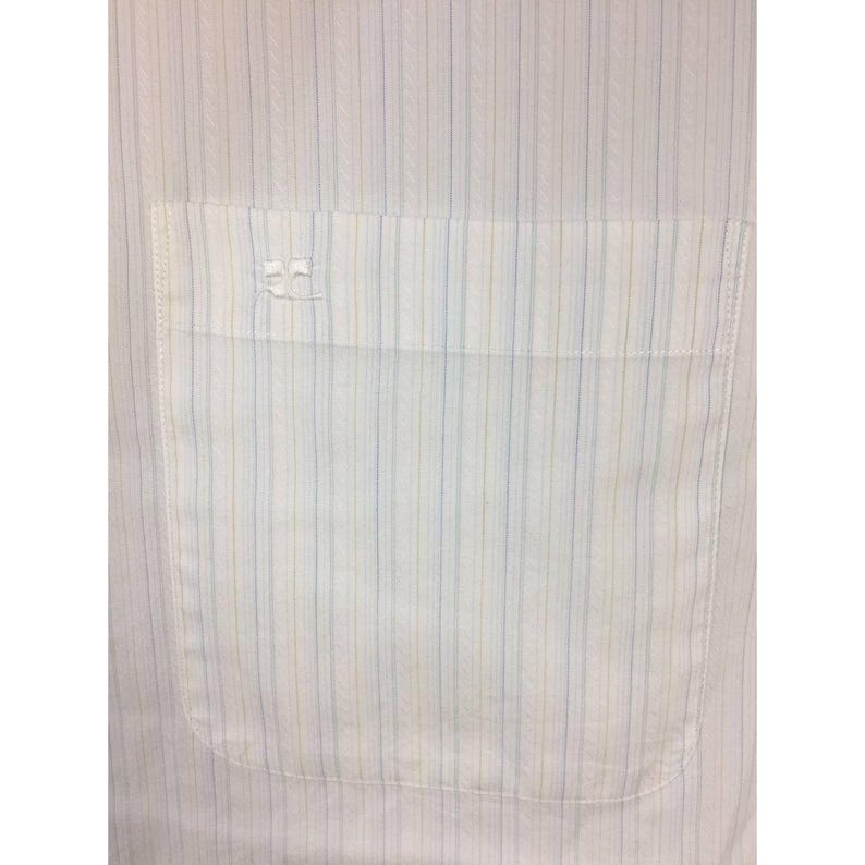 May include: A white button-down shirt featuring a subtle vertical pinstripe pattern in light blue and tan. The shirt includes a single chest pocket with a small, white embroidered logo detail positioned on the upper left corner of the pocket.
