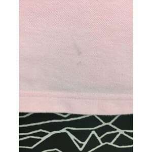 May include: Close-up of a light pink polo shirt. The fabric has a textured appearance. There are a few dark spots on the shirt. The bottom hem is visible, and the background is a black and white geometric pattern.