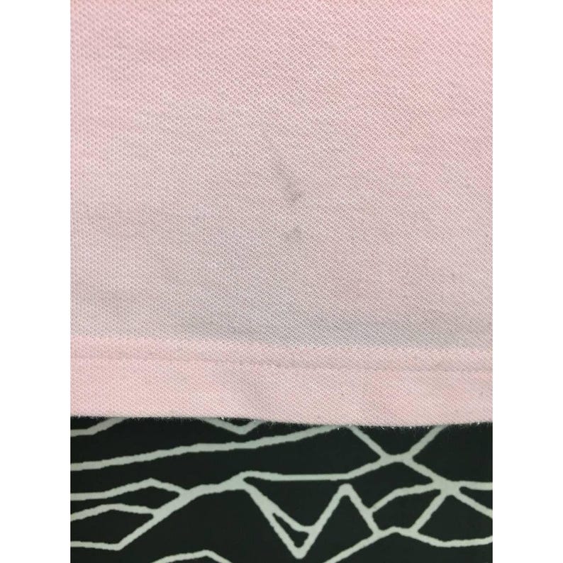 May include: Close-up of a light pink polo shirt. The fabric has a textured appearance. There are a few dark spots on the shirt. The bottom hem is visible, and the background is a black and white geometric pattern.