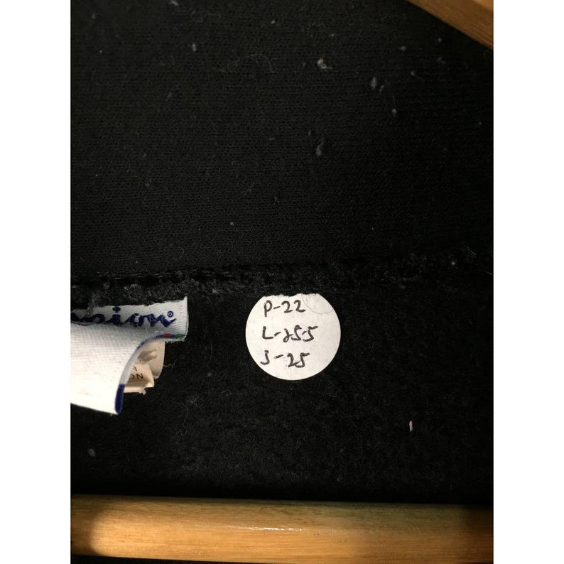 May include: Close-up of a black Champion sweatshirt. The image shows a white tag with the Champion logo and a circular sticker with handwritten text: P-22, L-355, and 3-25. The sweatshirt is hanging on a wooden hanger.