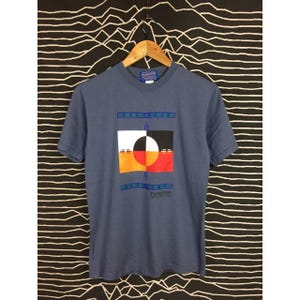 Könnte beinhalten: Ein blaues T-Shirt mit kurzen Ärmeln und einem geometrischen Design in Weiß, Schwarz, Orange und Rot. Das Shirt trägt den Markennamen "Pendleton". Das Shirt hängt an einem Holzbügel vor einem schwarz-weiß gemusterten Hintergrund.