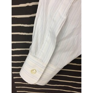 May include: A white long-sleeve dress shirt cuff featuring thin vertical pinstripes in light blue and tan. The cuff is secured with a single round, translucent white button. The fabric shows a crisp, structured weave with visible stitching.