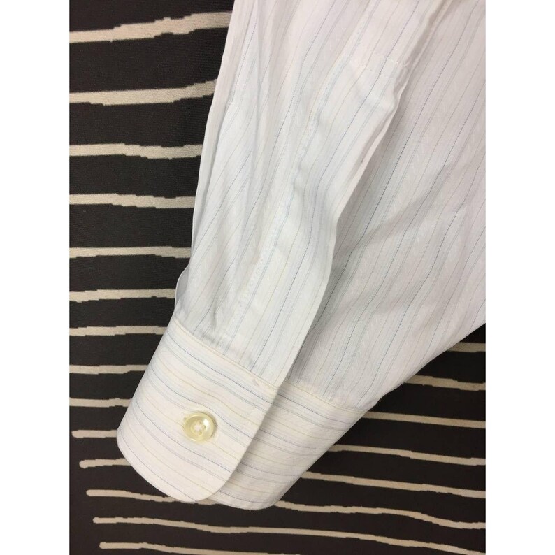 May include: A white long-sleeve dress shirt cuff featuring thin vertical pinstripes in light blue and tan. The cuff is secured with a single round, translucent white button. The fabric shows a crisp, structured weave with visible stitching.