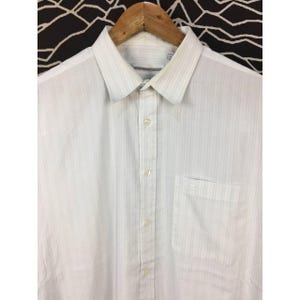 May include: A white button-down shirt featuring a subtle vertical pinstripe pattern in light blue and grey. The shirt includes a standard collar, a front button placket with light-colored buttons, and a single chest pocket on the left side.