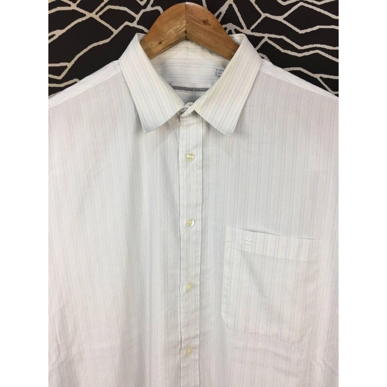May include: A white button-down shirt featuring a subtle vertical pinstripe pattern in light blue and grey. The shirt includes a standard collar, a front button placket with light-colored buttons, and a single chest pocket on the left side.