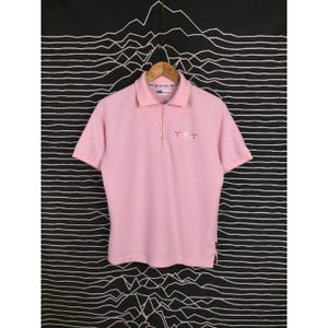 May include: A light pink polo shirt with short sleeves and a zipper closure. The shirt has a collared neckline and small embroidered details on the chest. The shirt is hanging on a wooden hanger against a black and white patterned background.