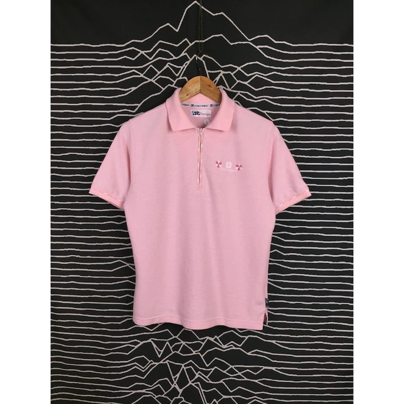 May include: A light pink polo shirt with short sleeves and a zipper closure. The shirt has a collared neckline and small embroidered details on the chest. The shirt is hanging on a wooden hanger against a black and white patterned background.