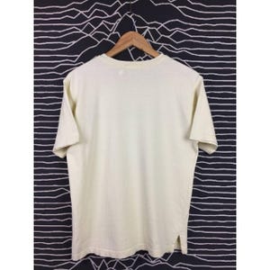Op de afbeelding: Cr&egrave;mekleurig T-shirt met korte mouwen aan een houten hanger. Het shirt heeft een eenvoudig ontwerp met een ronde hals en een rechte zoom. De stof lijkt een zacht, lichtgewicht materiaal te zijn. De achtergrond is een zwart-wit gestreept patroon.