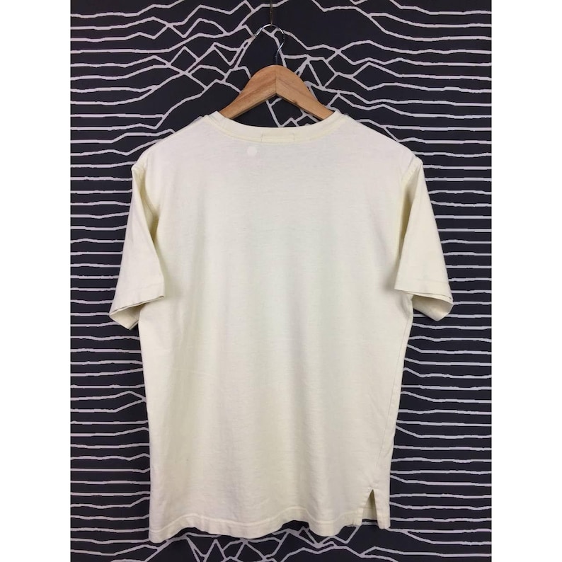 Op de afbeelding: Cr&egrave;mekleurig T-shirt met korte mouwen aan een houten hanger. Het shirt heeft een eenvoudig ontwerp met een ronde hals en een rechte zoom. De stof lijkt een zacht, lichtgewicht materiaal te zijn. De achtergrond is een zwart-wit gestreept patroon.