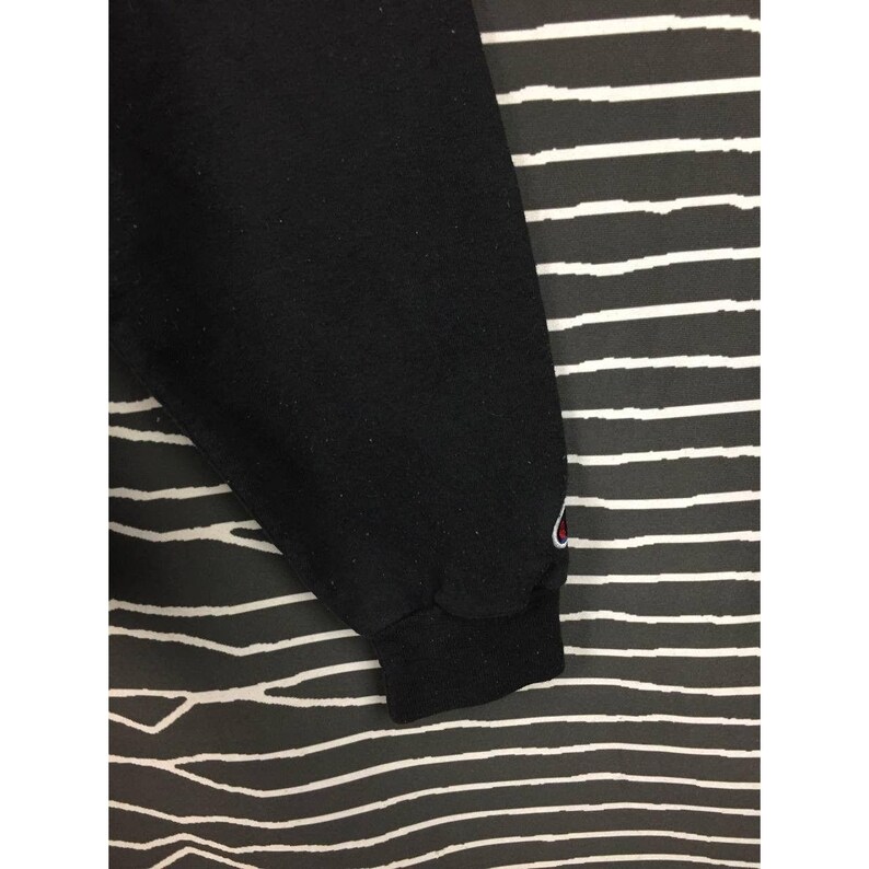 May include: Black sweatshirt sleeve with a ribbed cuff. A small red and white logo is visible near the cuff. The background is black and white striped.