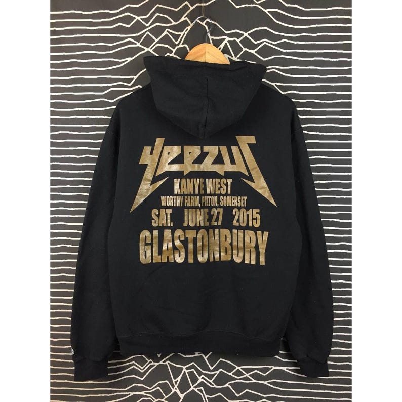 May include: Black hooded sweatshirt with gold lettering. The text on the back reads "YEEZUS" above "Kanye West" and "Glastonbury" with the date "SAT. JUNE 27 2015". The sweatshirt is hanging on a wooden hanger.