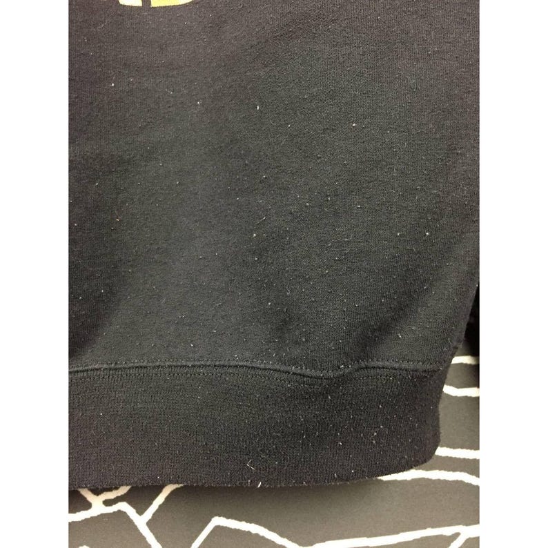 May include: Close-up of a black sweatshirt with a ribbed hem and cuffs. The fabric has a slightly worn appearance with small, light-colored flecks. The bottom hem is a darker shade of black.
