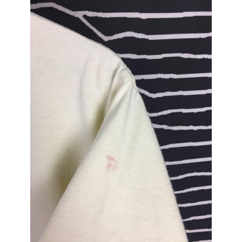 Op de afbeelding: Cr&egrave;mekleurige t-shirtmouw met een kleine roze vlek. De mouw is gericht op een zwart-wit gestreepte achtergrond.