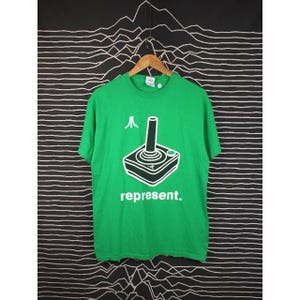 May include: Green t-shirt featuring a white joystick graphic and the word "represent." The Atari logo is above the joystick. The shirt is displayed on a wooden hanger against a black and white striped backdrop.