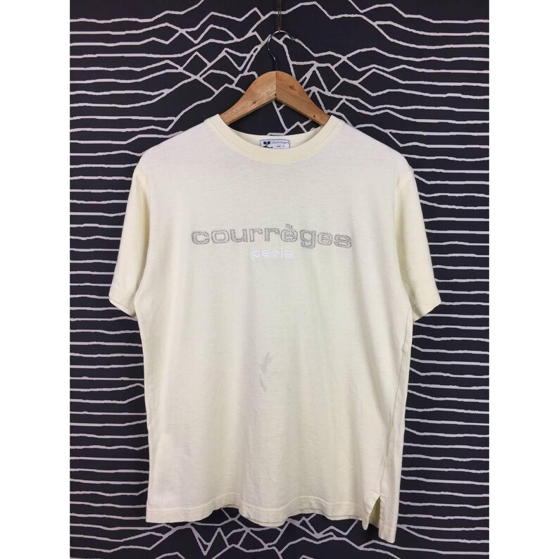 Op de afbeelding: Cr&egrave;mekleurig Courr&egrave;ges Paris T-shirt. De merknaam staat in een zilveren, omlijnde lettertype over de borst. Het shirt heeft korte mouwen en een ronde hals. Het shirt hangt aan een houten hanger.