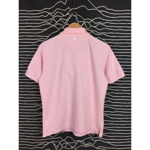 May include: A light pink polo shirt with short sleeves, a collared neckline, and a small logo on the back. The shirt is hanging on a wooden hanger against a black and white patterned background.