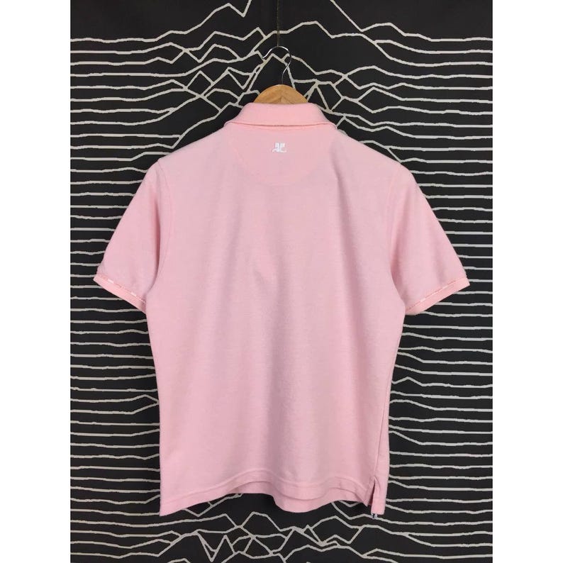 May include: A light pink polo shirt with short sleeves, a collared neckline, and a small logo on the back. The shirt is hanging on a wooden hanger against a black and white patterned background.