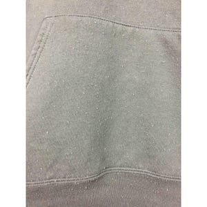 May include: Close-up of a gray hoodie pocket and fabric. The fabric has a textured appearance with small white specks scattered across the surface. The stitching is visible along the pocket edges and the bottom hem.