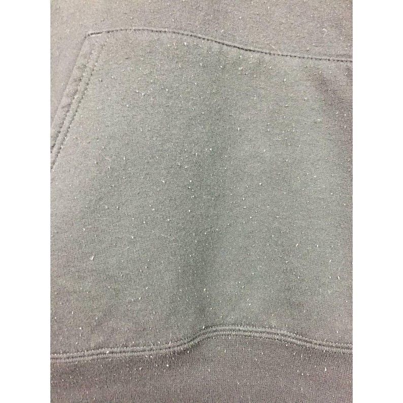 May include: Close-up of a gray hoodie pocket and fabric. The fabric has a textured appearance with small white specks scattered across the surface. The stitching is visible along the pocket edges and the bottom hem.