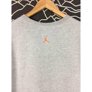 Peut inclure: Un t-shirt gris chin&eacute; &agrave; col rond avec un logo Jumpman orange brod&eacute; dans le haut du dos. Le t-shirt est suspendu &agrave; un cintre en bois sur un fond &agrave; motifs noir et blanc. Le t-shirt est fait d'une mati&egrave;re douce et confortable.