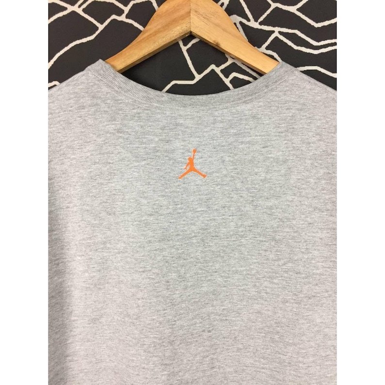 Peut inclure: Un t-shirt gris chin&eacute; &agrave; col rond avec un logo Jumpman orange brod&eacute; dans le haut du dos. Le t-shirt est suspendu &agrave; un cintre en bois sur un fond &agrave; motifs noir et blanc. Le t-shirt est fait d'une mati&egrave;re douce et confortable.