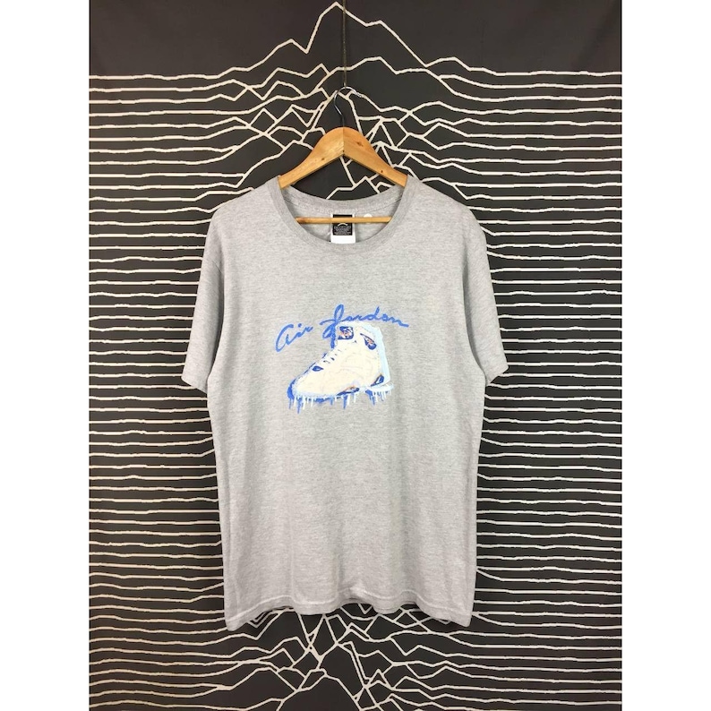 Peut inclure: T-shirt gris avec un motif de chaussure de basket-ball blanche et bleue et l'inscription "Air Jordan" en bleu. Le t-shirt est suspendu &agrave; un cintre en bois sur un fond &agrave; rayures noires et blanches.