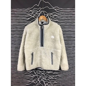 Puede incluir: Chaqueta de forro polar color crema con cuello alto y cierre de cremallera. La chaqueta presenta una cremallera azul marino y dos bolsillos laterales. Un pequeño logotipo blanco es visible en el pecho. La chaqueta está colgada en una percha de madera.