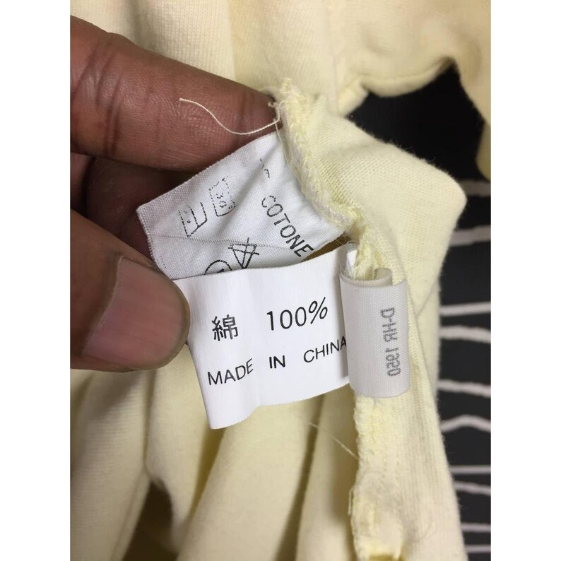 Op de afbeelding: Close-up van een lichtgeel kledinglabel. Op het label staat "COTTONE" en "100%" in het Japans en Engels. Op het label staat ook "MADE IN CHINA". Een klein wit label met "D-HB 1920" is ook zichtbaar.