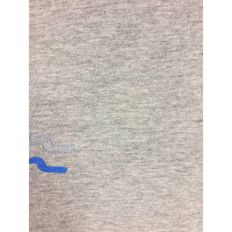 Peut inclure: Gros plan d'un t-shirt gris avec un motif brod&eacute; bleu. Le tissu a un aspect textur&eacute;. Le motif bleu est une forme stylis&eacute;e.
