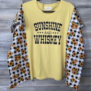 Può includere: Maglietta gialla a maniche lunghe con la scritta nera "SUNSHINE AND WHISKEY". Le maniche presentano un motivo a girasoli su sfondo a scacchi bianchi e neri. La maglietta è appesa a una gruccia nera.