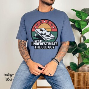 May include: A blue t-shirt with a vintage-style graphic of a cornhole game and the text "Never underestimate the old guy".