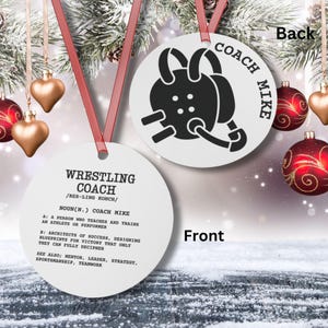 May include: A white round ornament with the words "Wrestling Coach" and a definition of the word "Coach". The back of the ornament has a black and white illustration of a wrestling headgear and the words "Coach Mike".