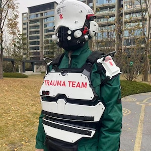 Puede incluir: Un casco blanco futurista y una armadura de pecho con detalles en rojo y la inscripción "TRAUMA TEAM" en rojo. El casco tiene una máscara negra y la armadura se lleva sobre un mono verde.