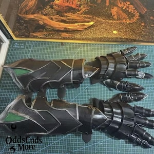 May include: A pair of black and silver gauntlets with articulated fingers. The gauntlets feature a segmented design with silver trim and green accents. The image shows the gauntlets on a green cutting mat. The logo "OddsEnds More" is visible.