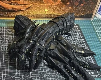 May include: A pair of black, articulated gauntlets with a textured, scale-like appearance. The gauntlets feature pointed extensions at the fingertips and are adorned with small, metallic studs. The design suggests a fantasy or cosplay theme.