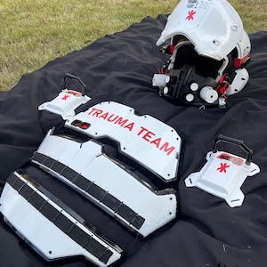 Puede incluir: Un conjunto de casco y armadura blancos y negros con detalles en rojo, con el texto "TRAUMA TEAM" en rojo. El conjunto incluye un casco, piezas para el pecho y los brazos, y dos pequeñas cajas, todas con un símbolo médico rojo.