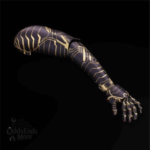 Puede incluir: Un brazo protésico negro con diseños dorados en forma de circuitos. El brazo está articulado en el codo y la muñeca, y la mano está relajada. El producto es de OddsEndsMore.