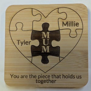 May include: A square wooden coaster with a heart-shaped puzzle design. The puzzle pieces feature the names "Tyler" and "Millie", with "MUM" in the center. The text "You are the piece that holds us together" is at the bottom.