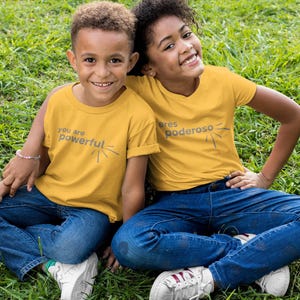 May include: Two children wearing yellow t-shirts with empowering messages in English and Spanish. The girl's shirt says "Eres Poderoso" and the boy's shirt says "You are Powerful". They are sitting on a green grassy surface.