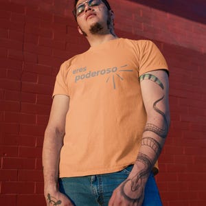 May include: A person wearing a light orange t-shirt with the text "eres poderoso" and a snake tattoo on their arm. The person is standing in front of a red brick wall.